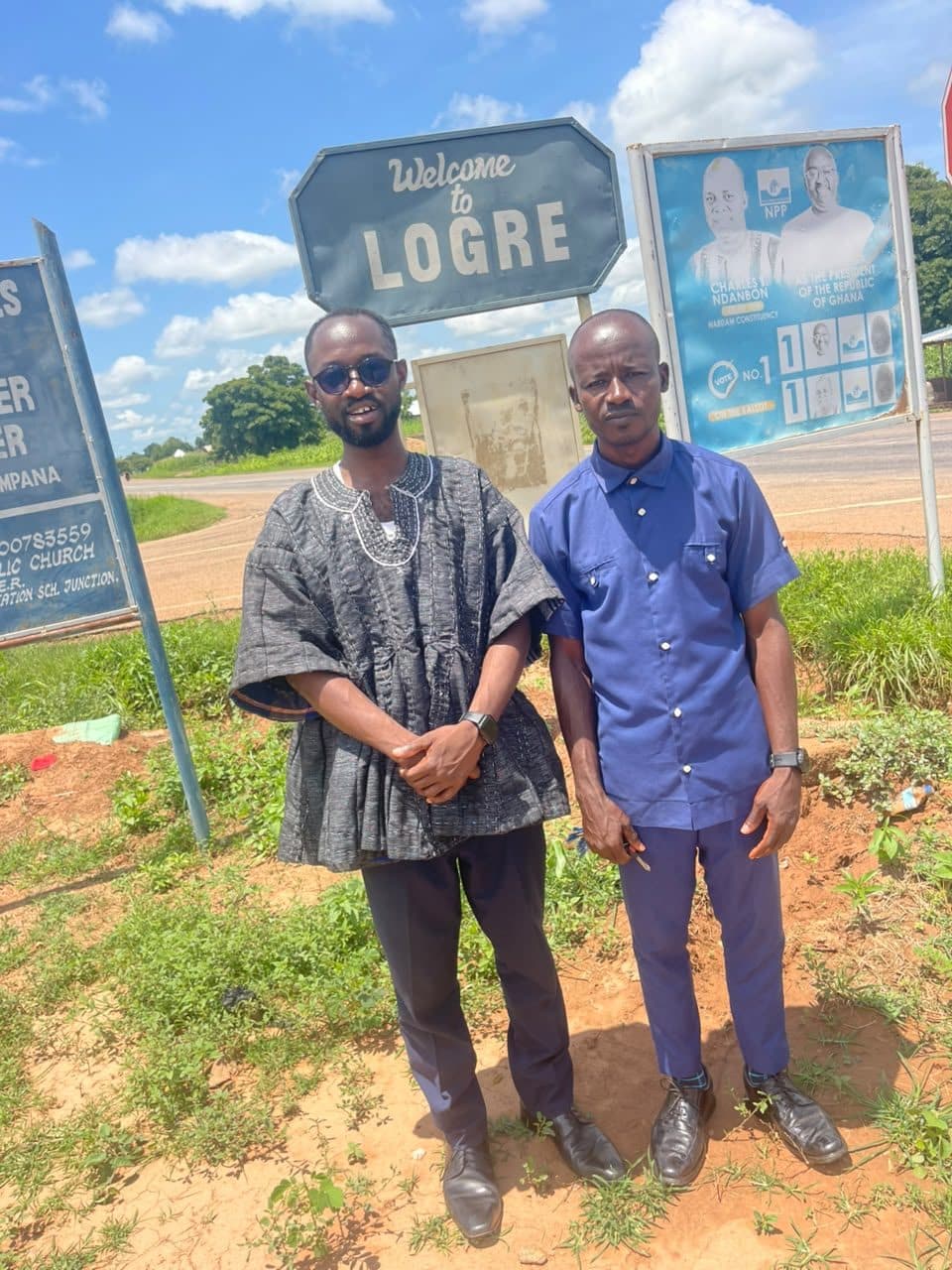 Patrick Attankurugu and Martin Asambo Atemgu at the Welcome to Logre signboard on the trunk road, September 2026